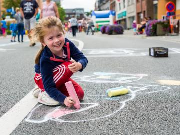 Een kind tekent met stoepkrijt op de weg en kijkt lachend op.